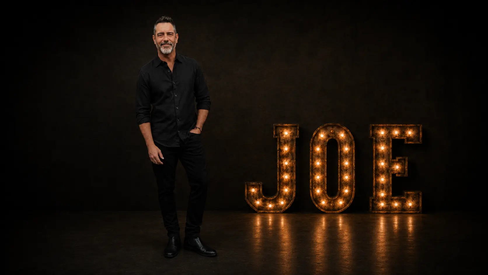 Joe Graham standing in a dark studio with illuminated JOE marquee letters.